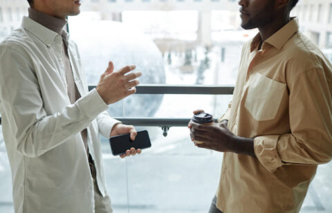 Cropped side view shot of two colleagues chatting standing against window and gesturing copy space